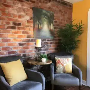A cozy corner with a painted faux brick accent wall in warm terracotta tones, featuring a pathway through a forest on a canvas. The scene is completed with two gray armchairs with yellow and patterned throw pillows, a round wooden side table, a decorative lamp, and a potted plant, creating a peaceful and inviting nook.