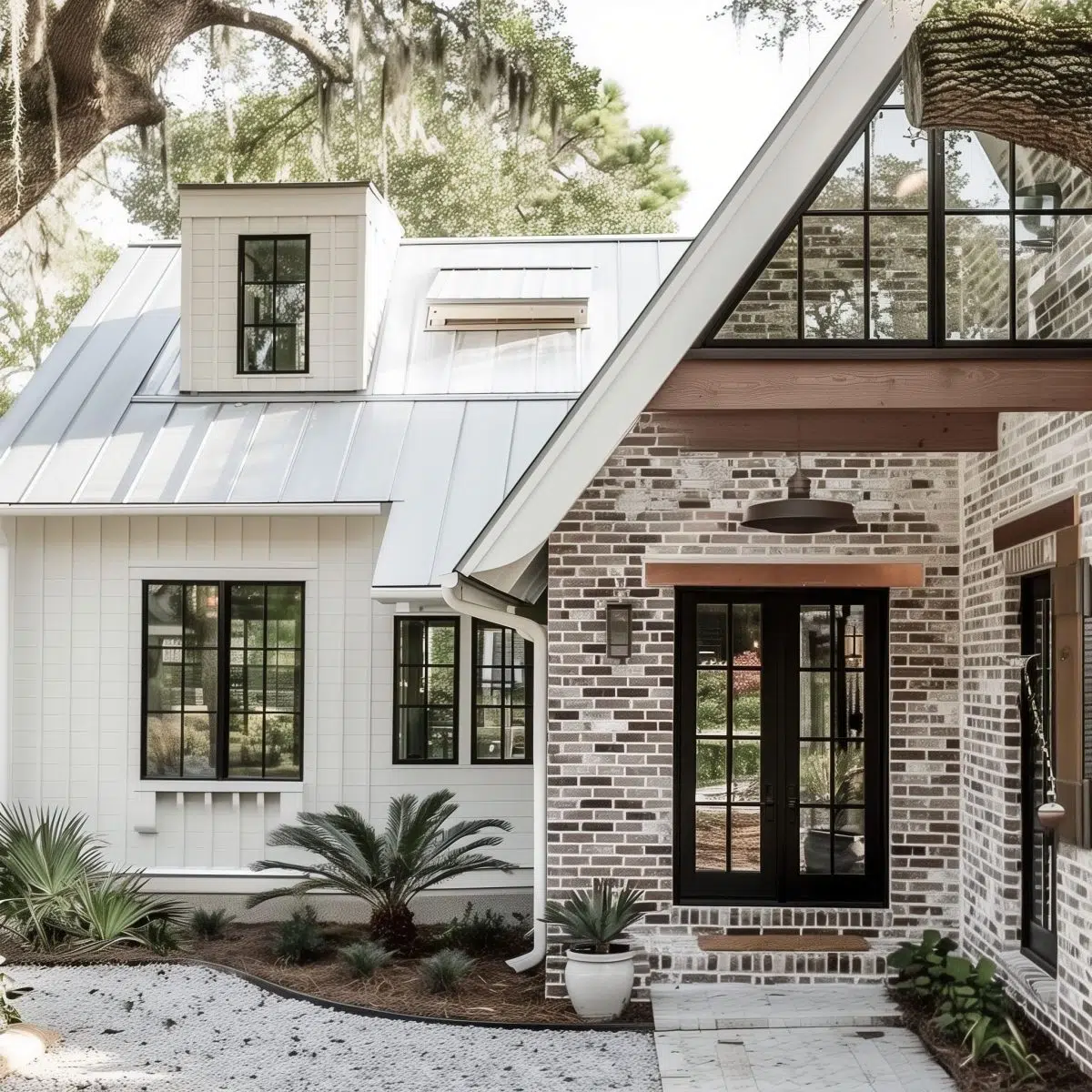 A stylish house with a coastal vibe, combining white vertical siding and traditional red brick. The house has large black-framed windows, a metal roof, and is surrounded by sandy landscaping with small palm trees.