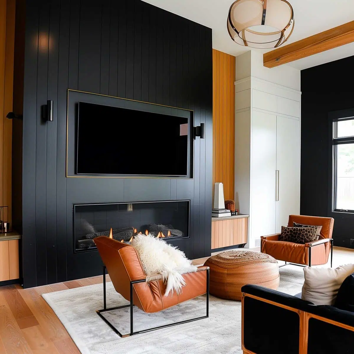 black_fireplace_wall (14) A modern living room with a black feature wall and a wall-mounted TV above a sleek fireplace. There are elegant leather armchairs with white fur throws, a round ottoman, and light wooden flooring. Large windows add natural light, and a unique chandelier hangs overhead.