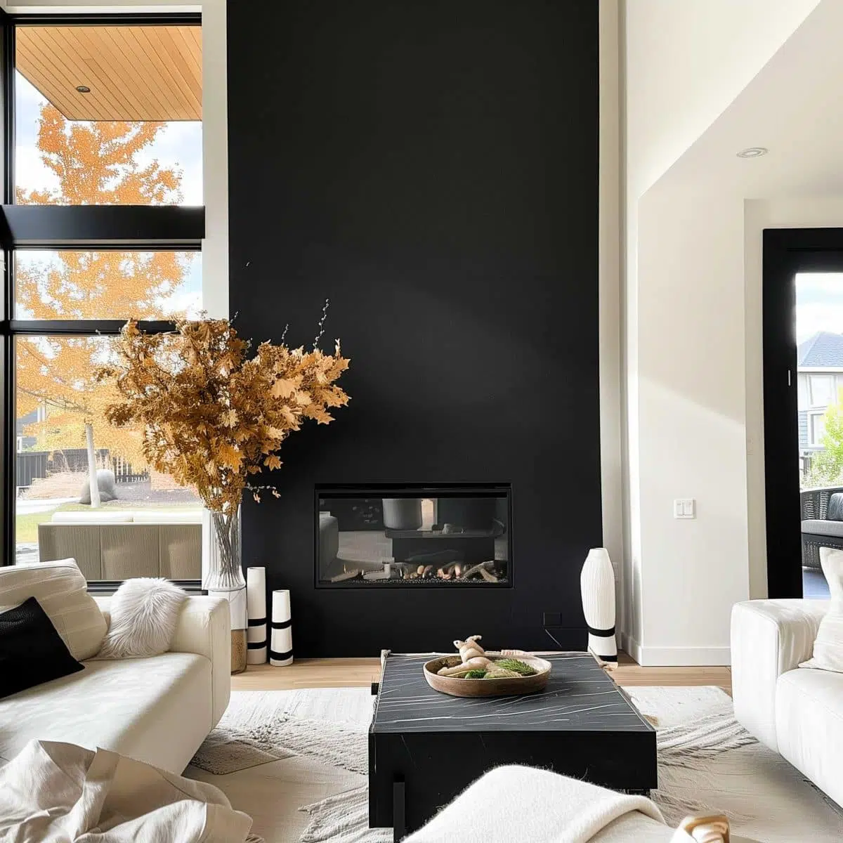 black_fireplace_wall (19) A modern living room features a black accent wall with a built-in fireplace, surrounded by white furniture. A black coffee table with decorative items sits in the center. A vase with dried leaves is near the window, letting in natural light and views of autumn foliage.