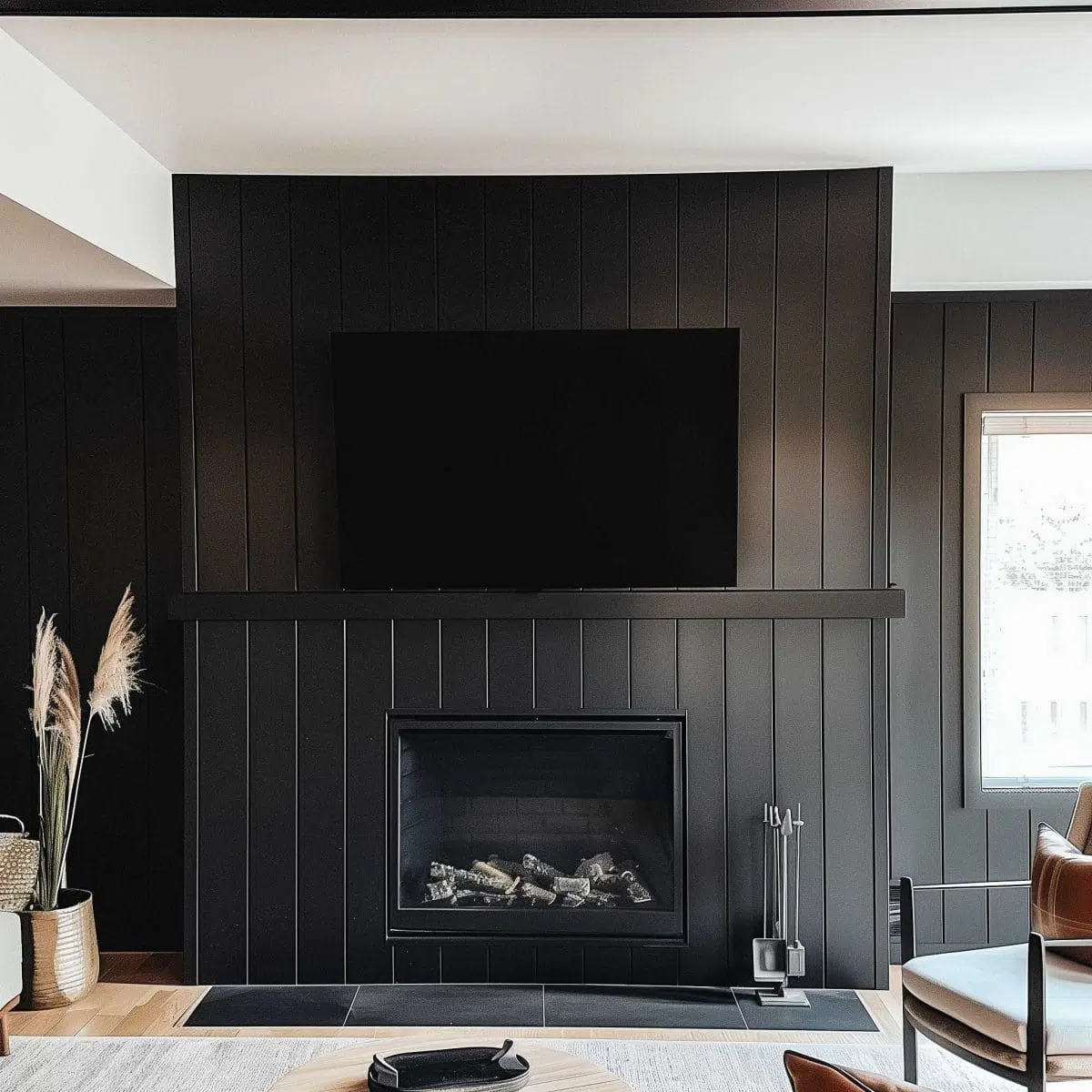 black_fireplace_wall (23) A modern living room features a sleek black fireplace wall with vertical paneling, a mounted flat-screen TV above the fireplace, and a log set in the firebox. Decorative elements include pampas grass in a vase on the left and a window on the right allowing natural light.