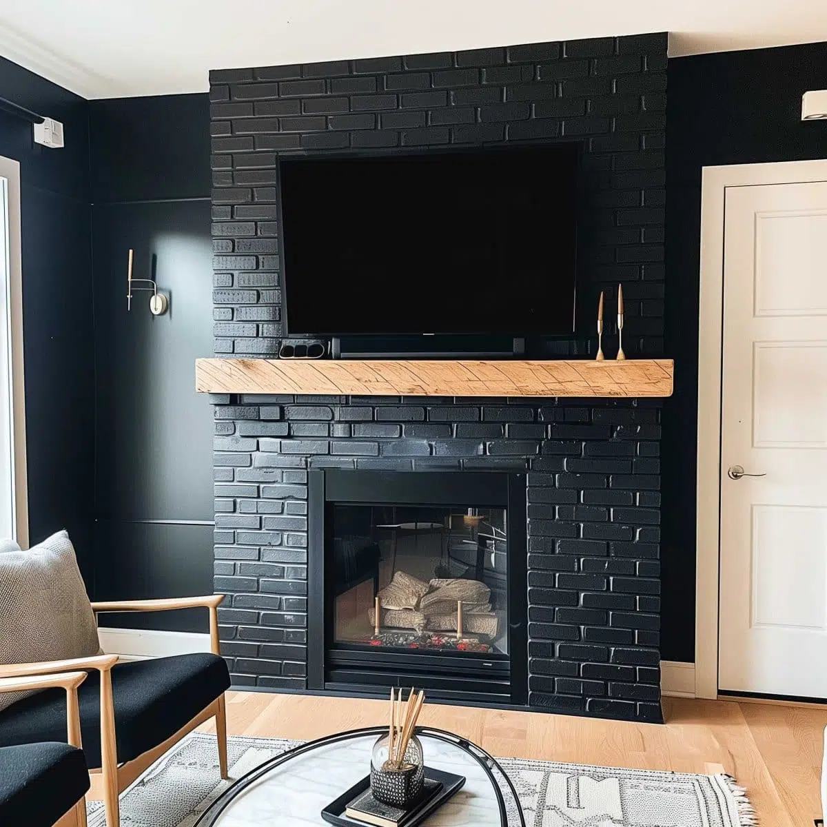 black_fireplace_wall (4) A modern living room featuring a black brick fireplace with a wooden mantel. A flat-screen TV is mounted above the mantel, and minimalist décor adorns the space. There is a black chair with light wood accents, a light-colored rug, and a coffee table in front.